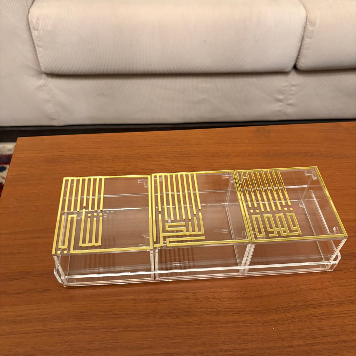 A three-compartment acrylic organizer box with gold-colored geometric patterns on the lids, placed on a wooden table with a sofa in the background.