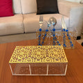 A gold laser-cut metal table runner with a geometric floral pattern sits on a wooden table in front of a clear acrylic storage box. A matching gold and clear utensil holder with blue handles and silver cutlery is positioned behind it on a sofa table, with a beige couch in the background.