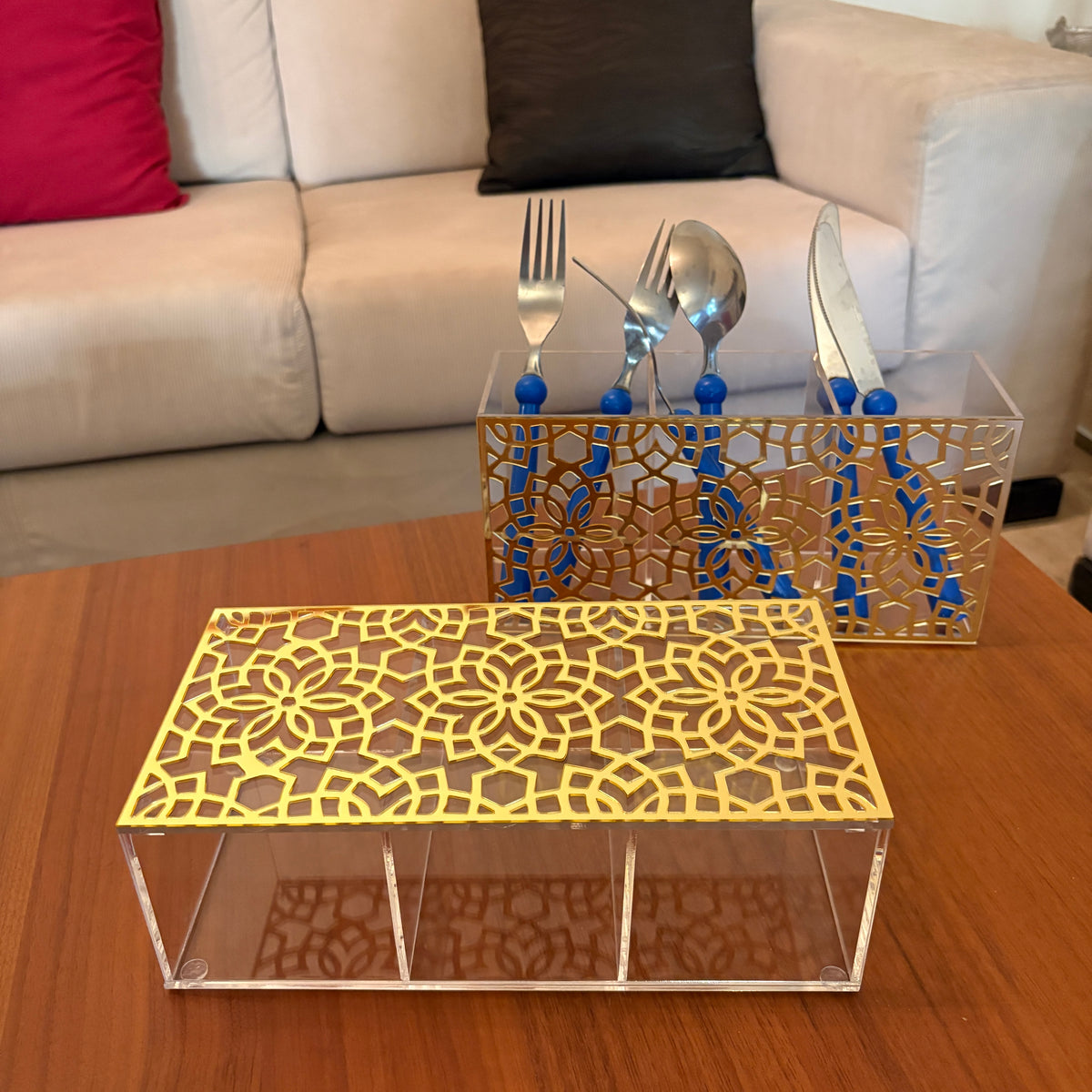 A gold laser-cut metal table runner with a geometric floral pattern sits on a wooden table in front of a clear acrylic storage box. A matching gold and clear utensil holder with blue handles and silver cutlery is positioned behind it on a sofa table, with a beige couch in the background.