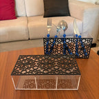 A black plexi cutout organizer tray with a clear acrylic base sits on a wooden table in front of a sofa. A matching set with blue-handled utensils is placed behind it.