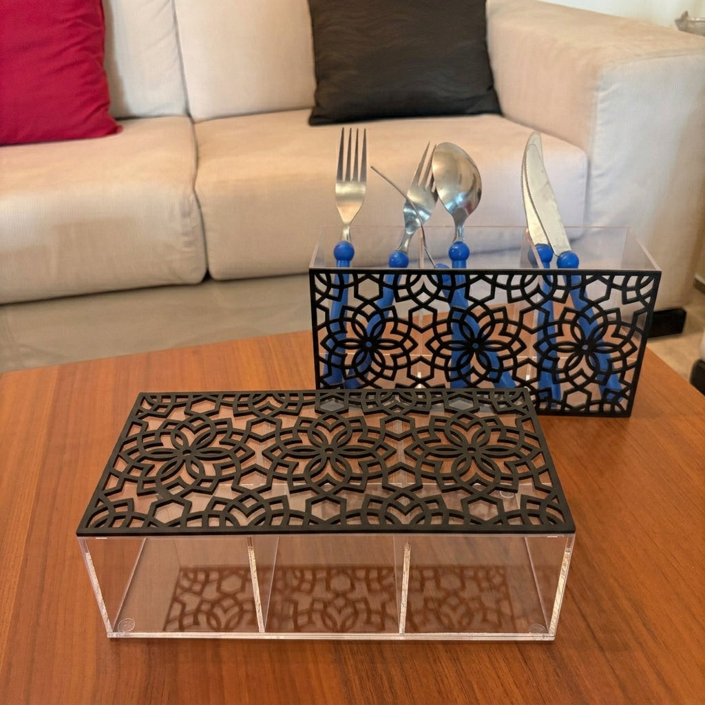 A black plexi cutout organizer tray with a clear acrylic base sits on a wooden table in front of a sofa. A matching set with blue-handled utensils is placed behind it.