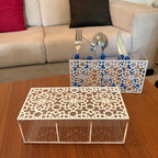 Two white laser-cut acrylic organizer trays with floral patterns, one holding silver cutlery in blue holders, displayed on a wooden table in front of a beige sofa.