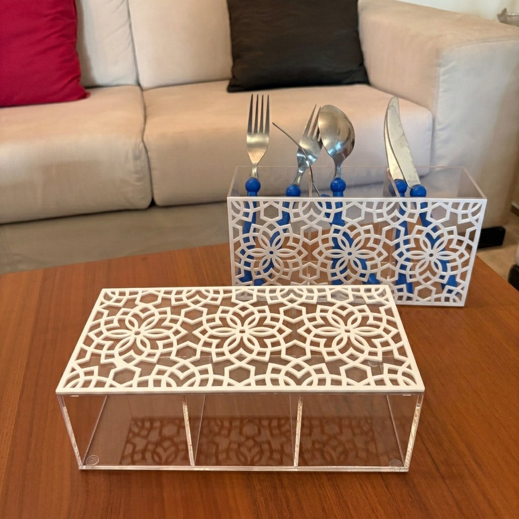 Two white laser-cut acrylic organizer trays with floral patterns, one holding silver cutlery in blue holders, displayed on a wooden table in front of a beige sofa.